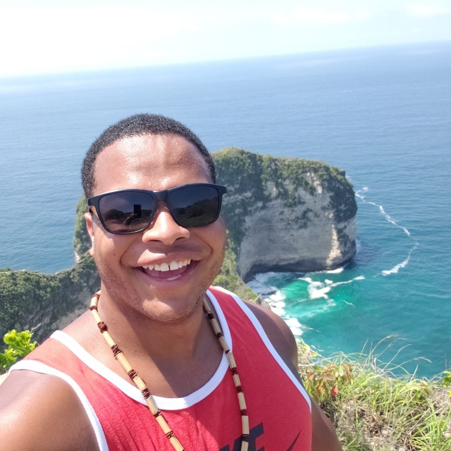 A man wearing sunglasses and a red tank top smiles in front of the ocean