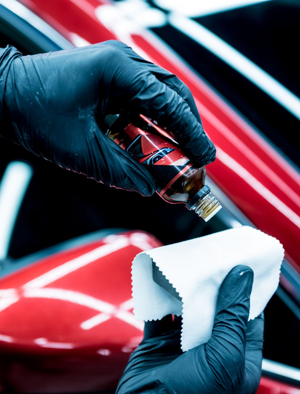 Black-gloved hands applying liquid from a bottle onto a white cloth in front of a red car.