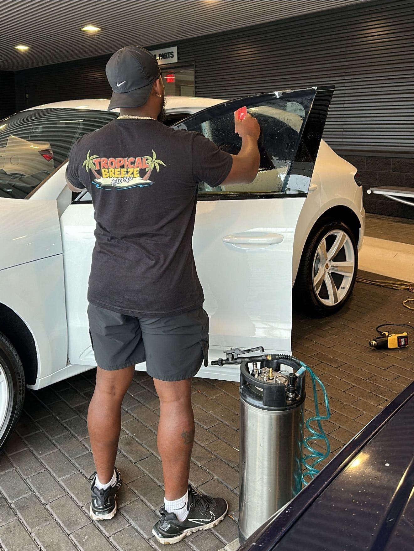 Person applying window tint to a white car. They wear a black shirt, shorts, and hat. Tinting equipment is nearby.