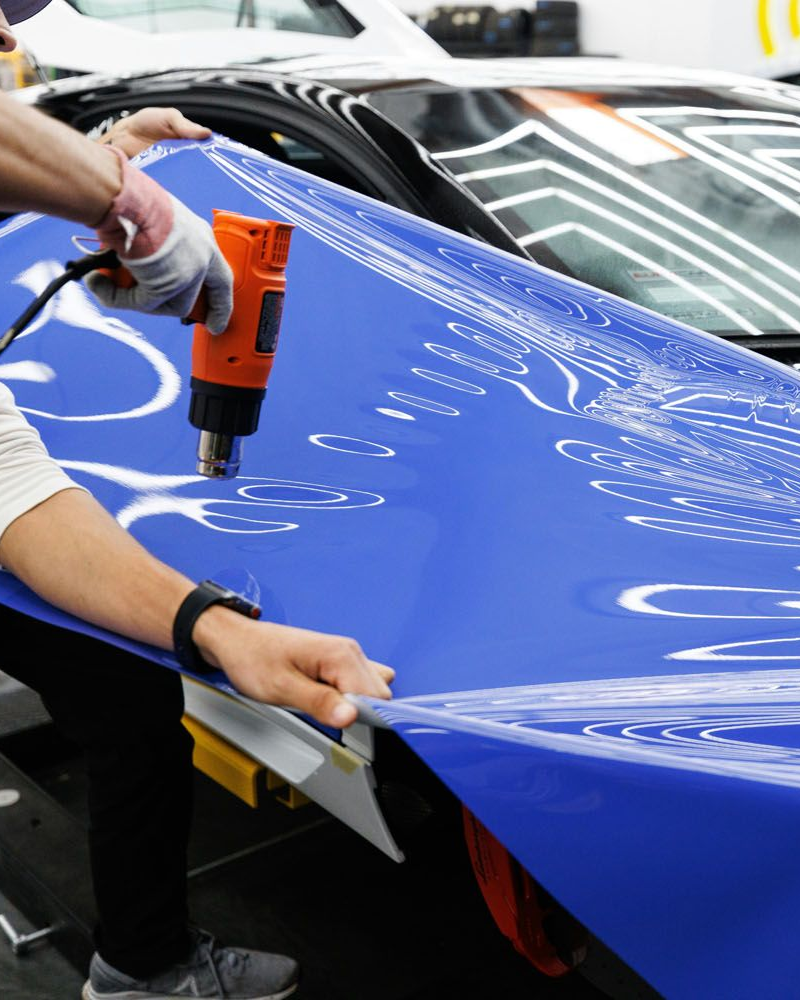 Person applying blue vinyl wrap to a car using a heat gun.