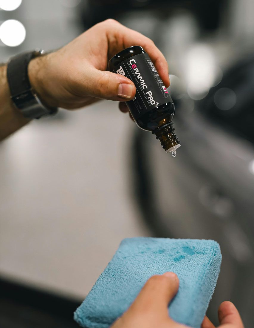 Person pouring liquid from a bottle onto a blue applicator pad, likely for car detailing.
