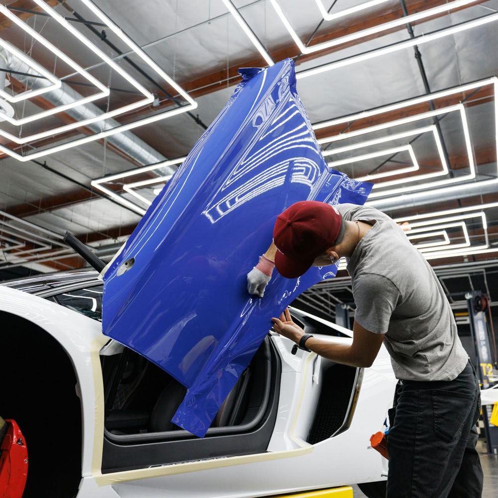 Person applying blue vinyl wrap to a white car door in a well-lit workshop.