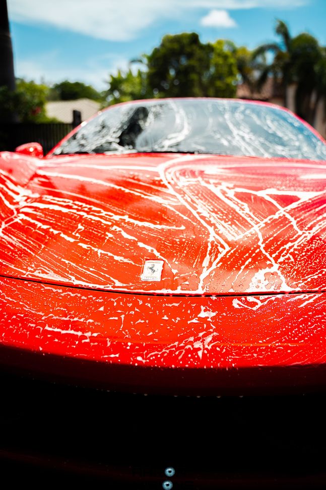 Red Ferrari car being washed with soap, outdoors on a sunny day.