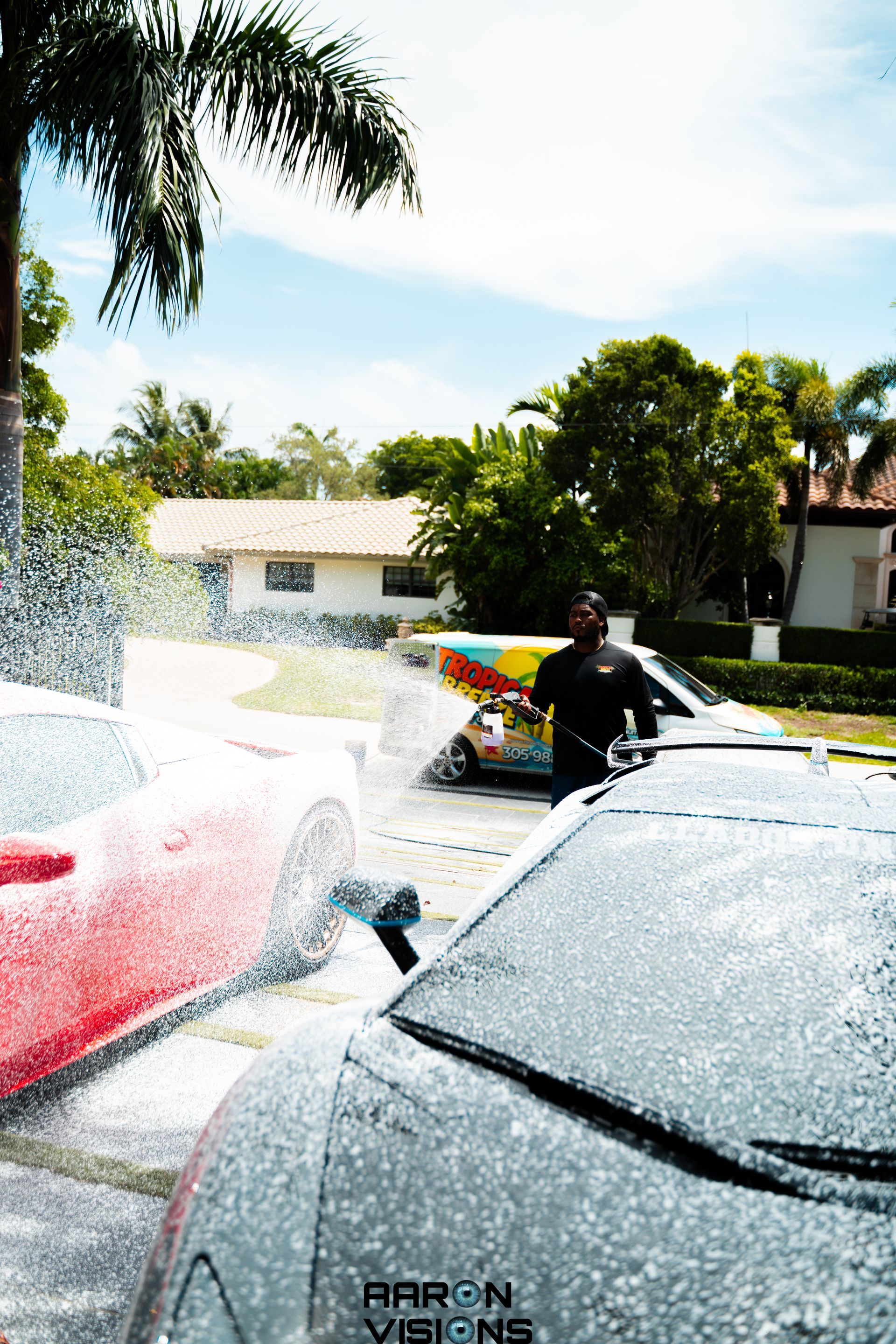 Man washing two cars with foamy soap in a driveway on a sunny day.