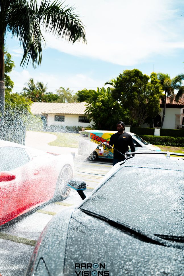 Man washing two cars with foamy soap in a driveway on a sunny day.