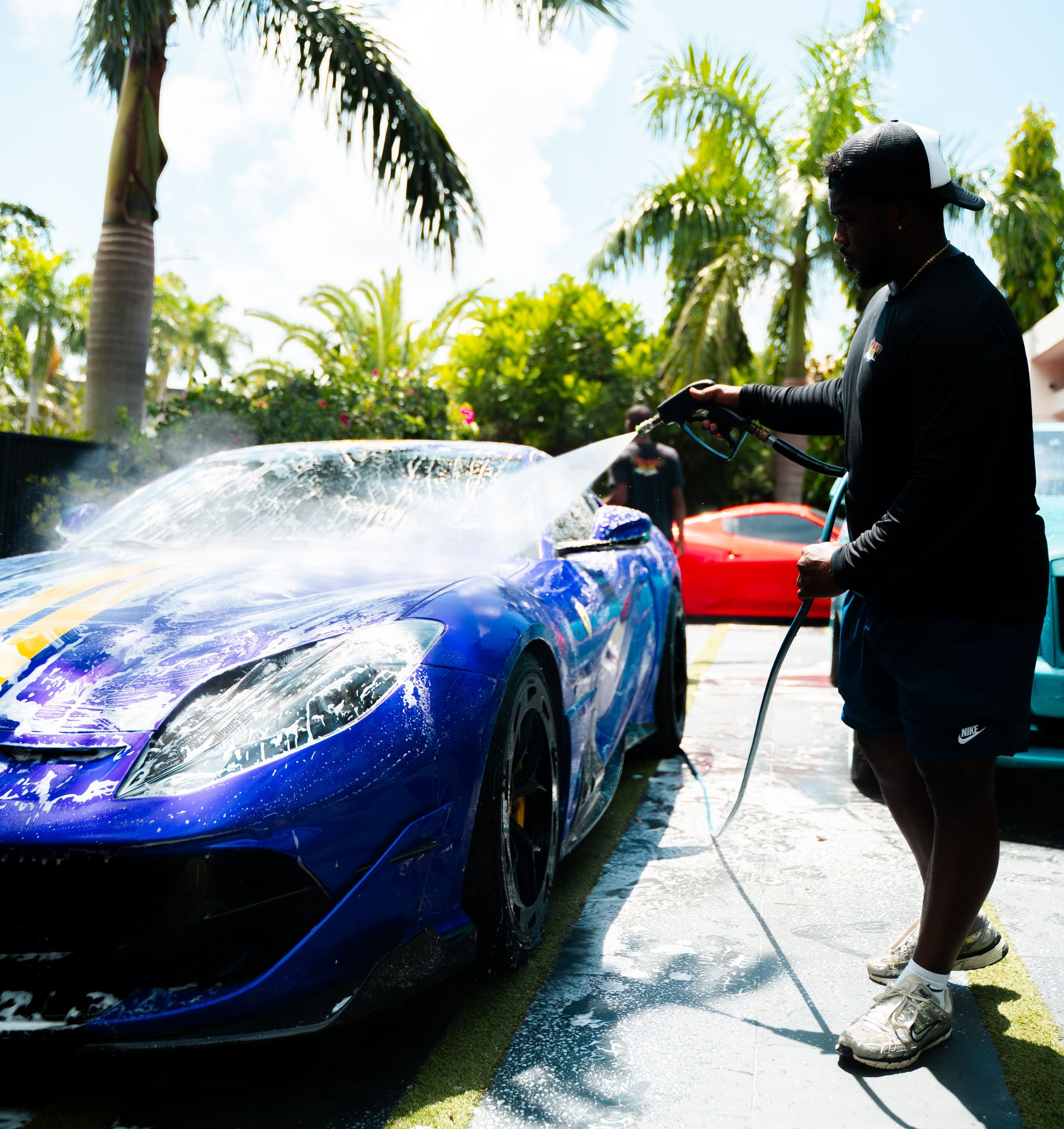 Person washing a blue sports car with yellow stripes using a hose. Other cars and palm trees are in the background.