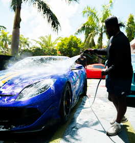 Person washing a blue sports car with yellow stripes using a hose. Other cars and palm trees are in the background.