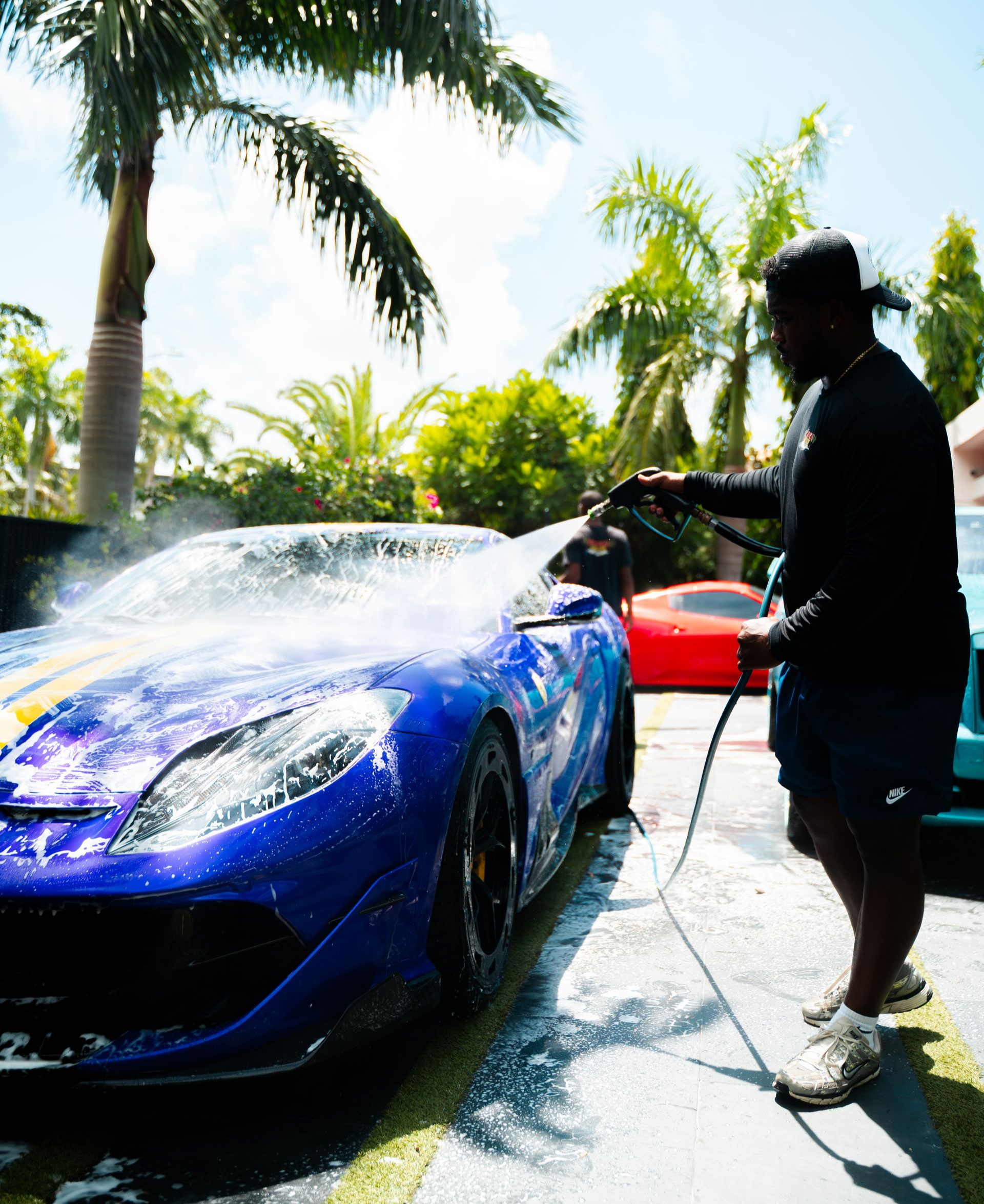 Person washes a blue sports car with yellow stripes using a hose outdoors.