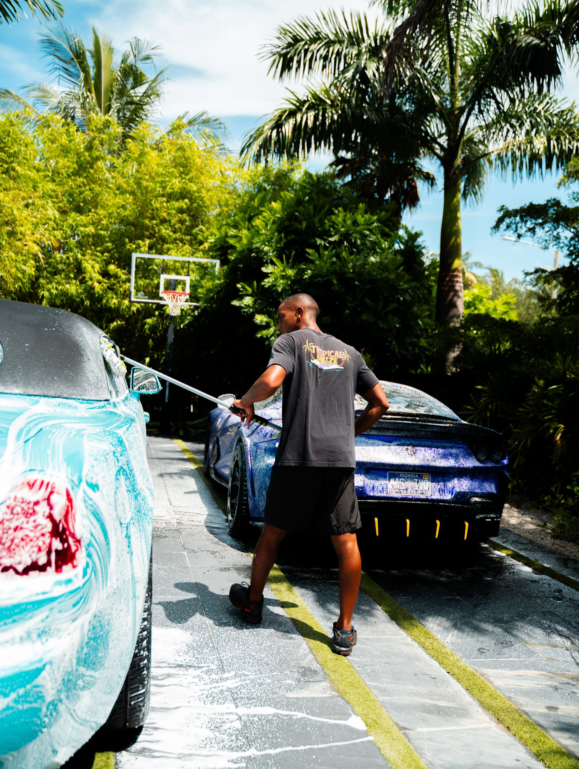 Man washing a blue sports car outside, basketball hoop in background.