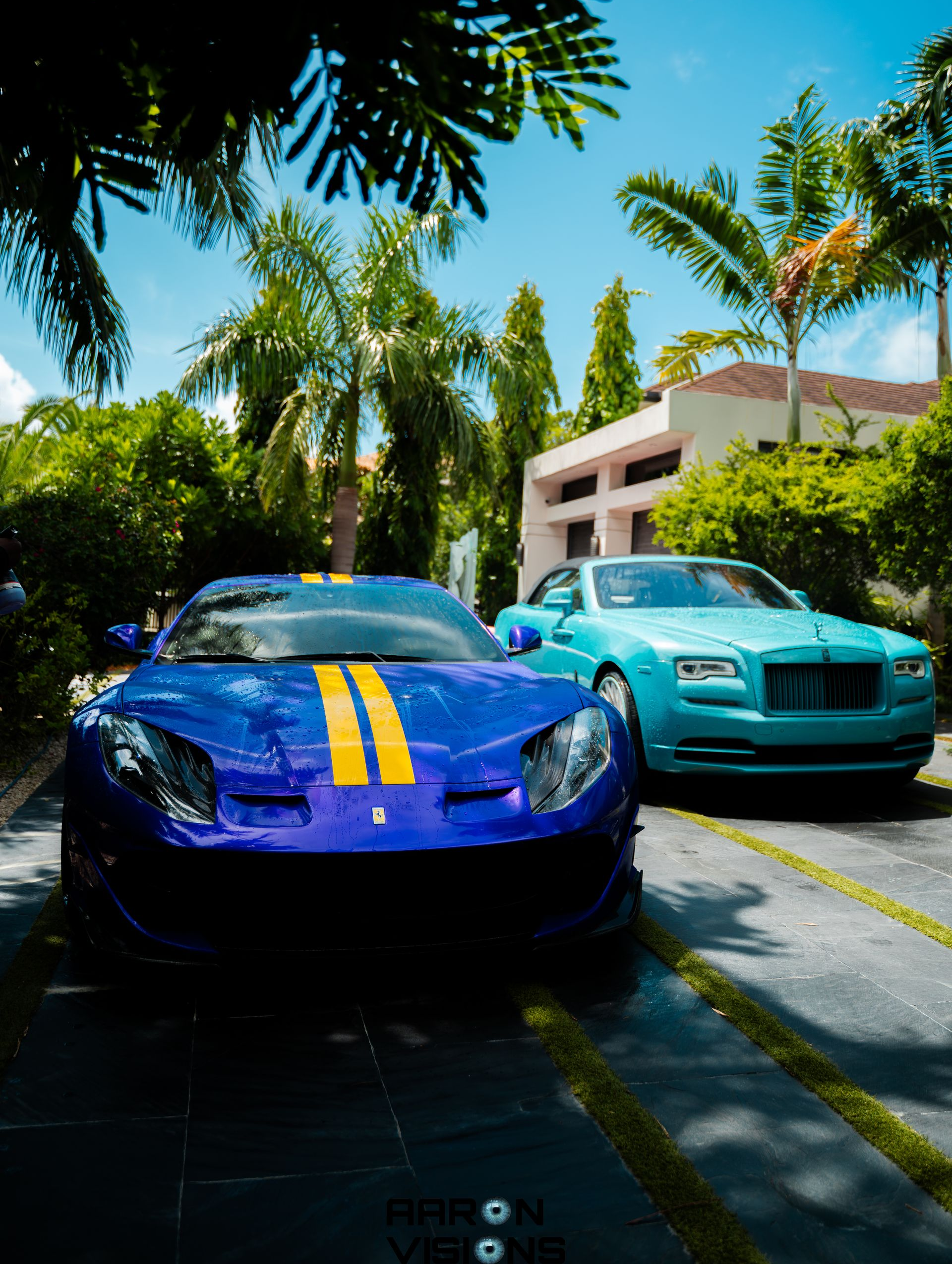 Blue Ferrari and turquoise Rolls Royce parked on a driveway with palm trees and a building in the background.