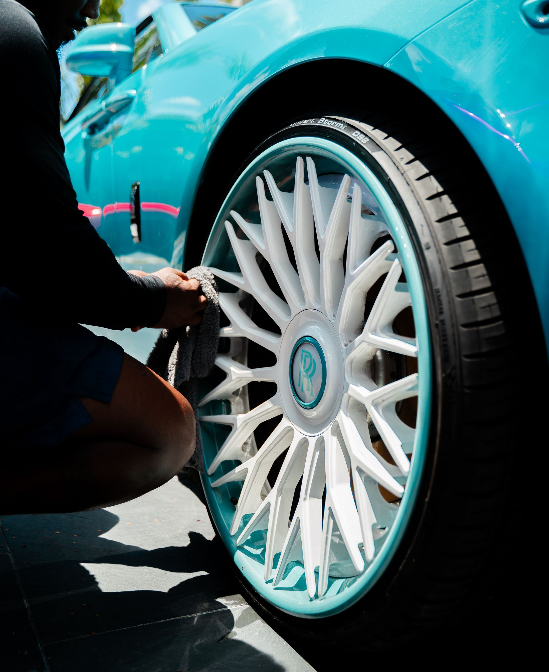 Person cleaning a teal car wheel with intricate white spokes.