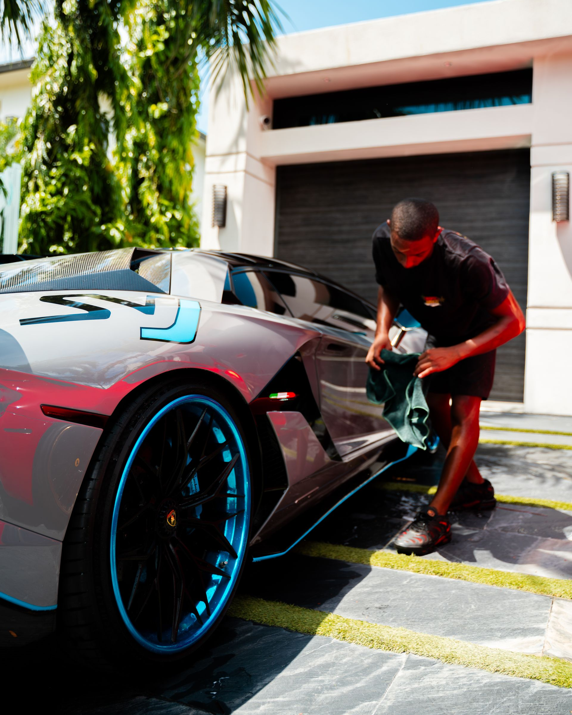 Person wiping down a silver Lamborghini with blue accents parked outside a modern house with a garage.