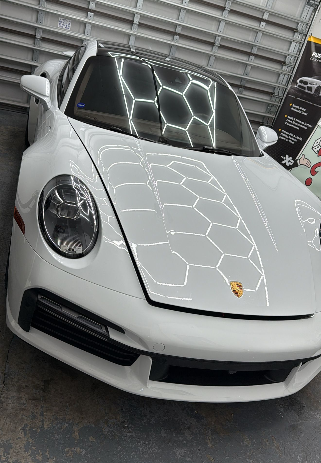 White Porsche sports car in a garage, reflecting hexagonal lights on its hood and roof.