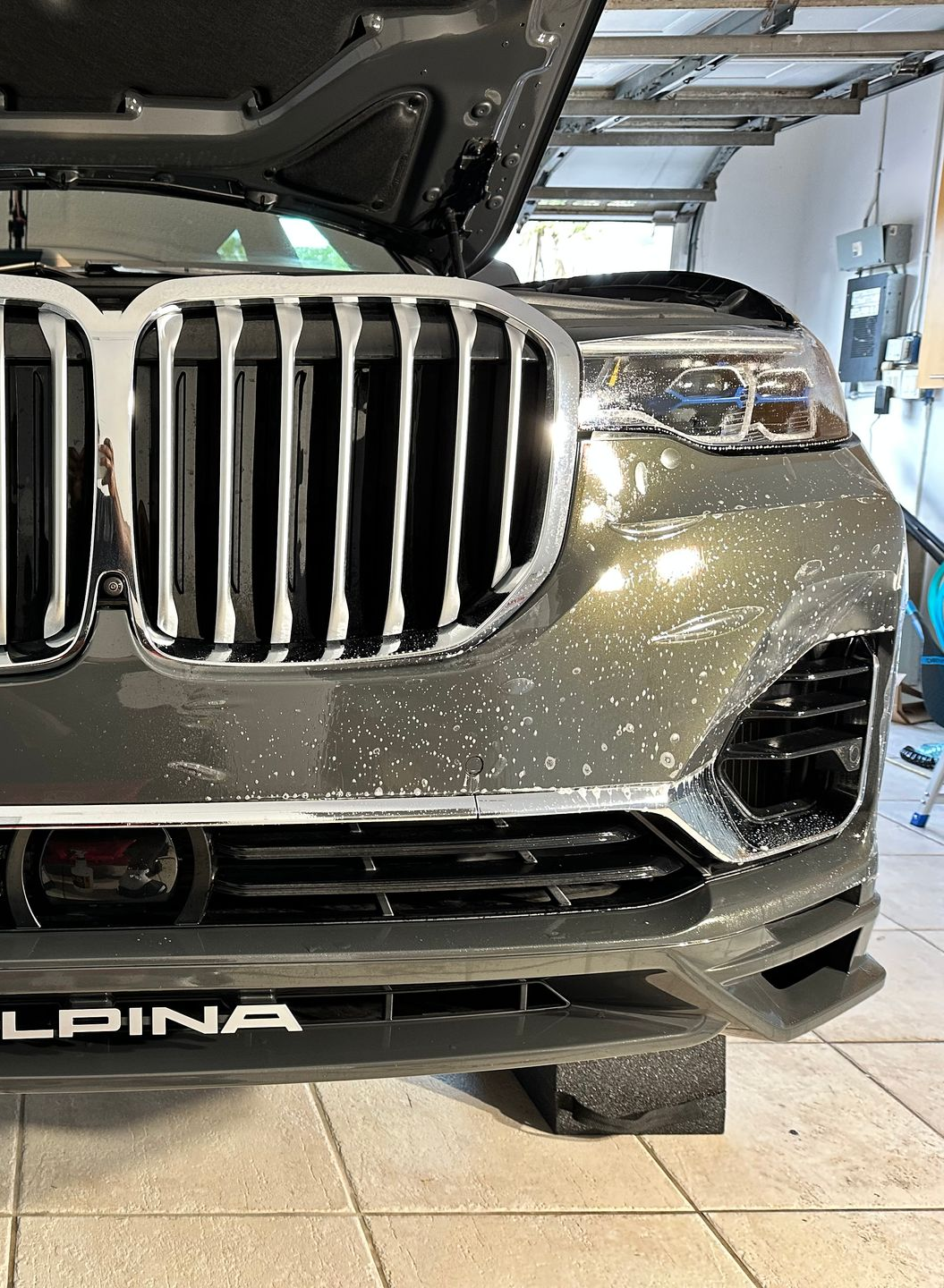Silver BMW Alpina car with open hood, covered in suds, parked in a garage.