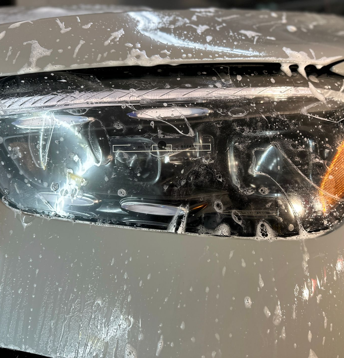 A car headlight covered in soapy water, partially obscured by gray film.