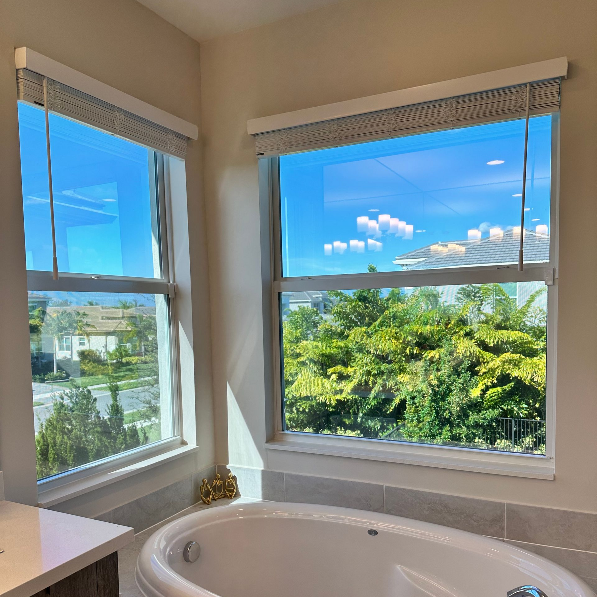 Two open windows in a bathroom offer views of a neighborhood and trees, above a white bathtub.