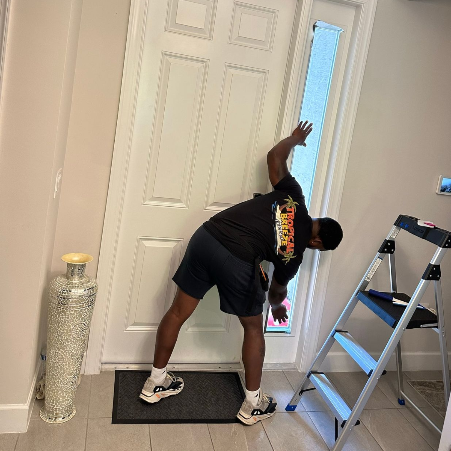 Person leans toward a white door, arm raised. A step ladder and decorative vase are nearby.