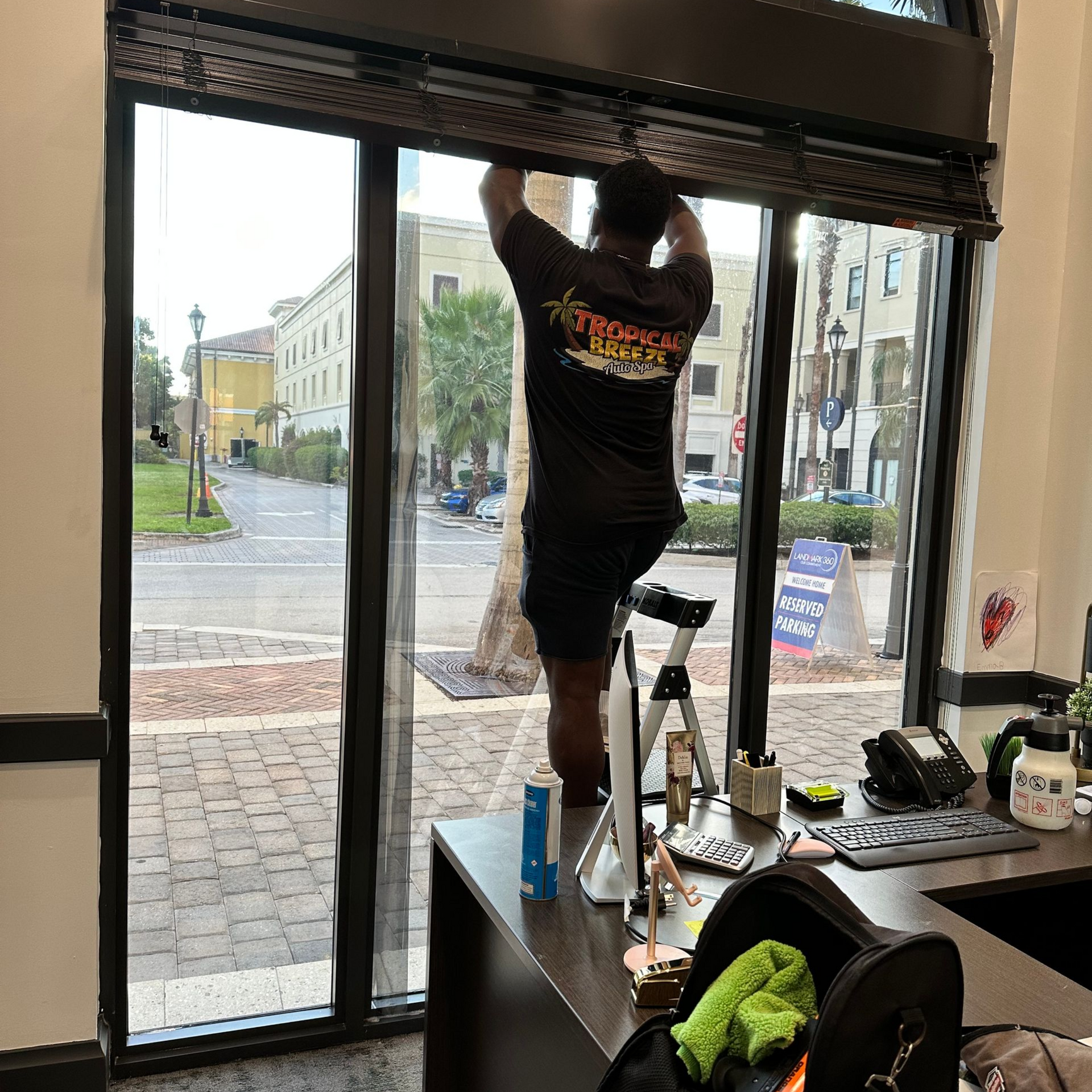 A person in a black shirt is on a step stool, adjusting a window blind in an office setting.
