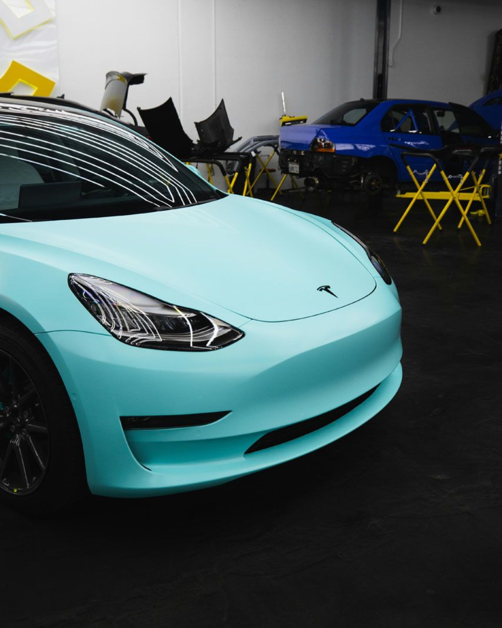 A light blue Tesla Model 3 in a garage, another blue car in the background.