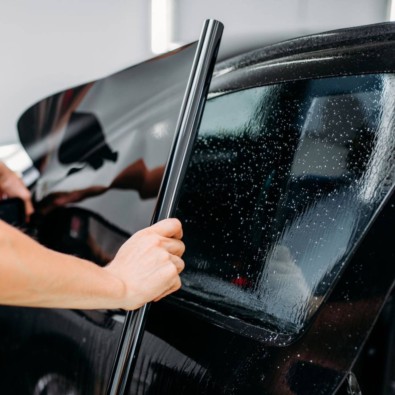 Person applying black tint to a car window, using a tool to smooth the film.