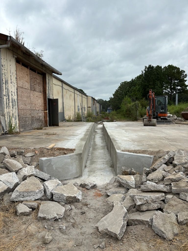 Concrete pit and demolition debris in front of a long industrial building, excavator at the end.