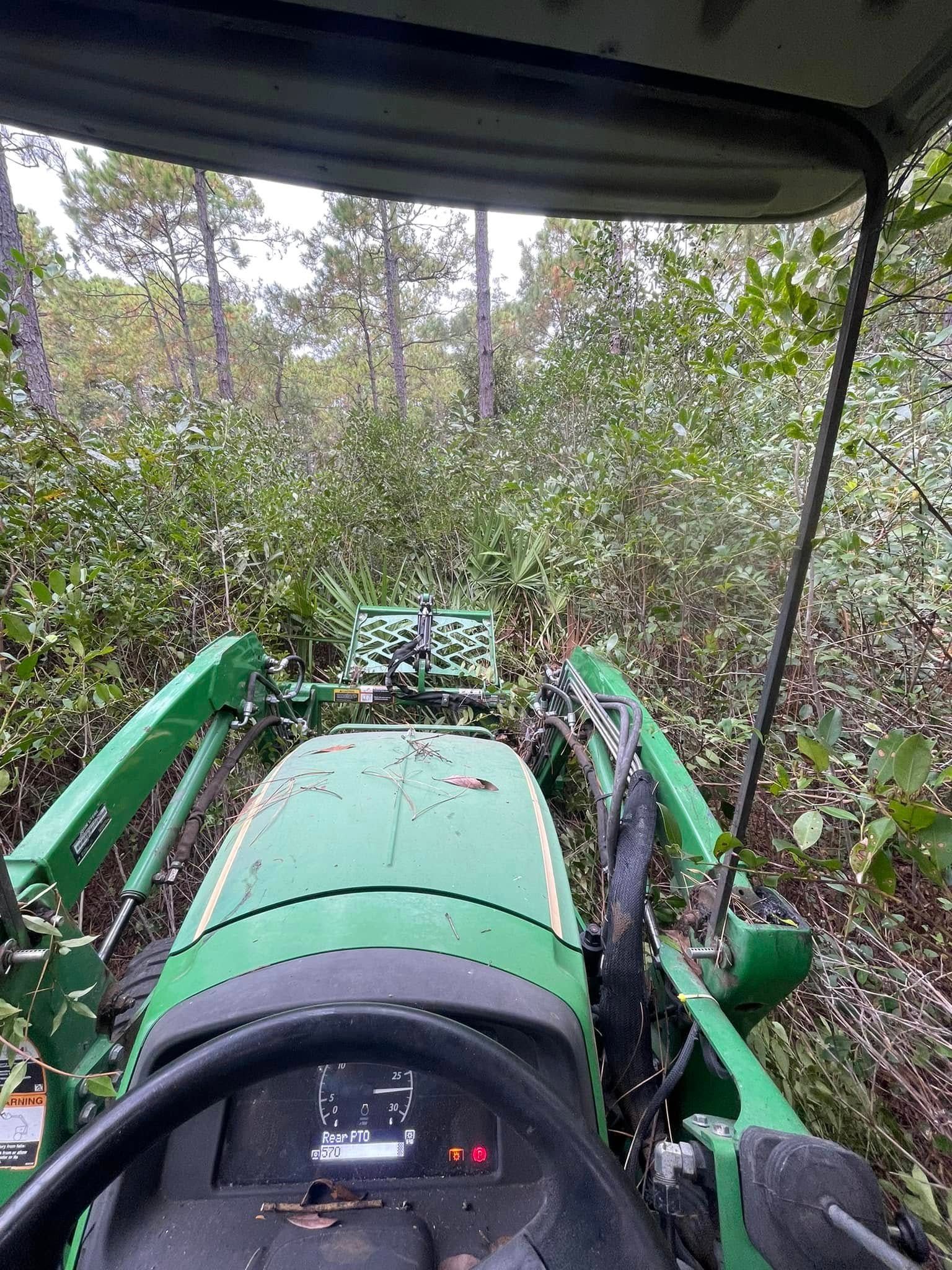 A person operating a green John Deere tractor clearing brush in a wooded area.