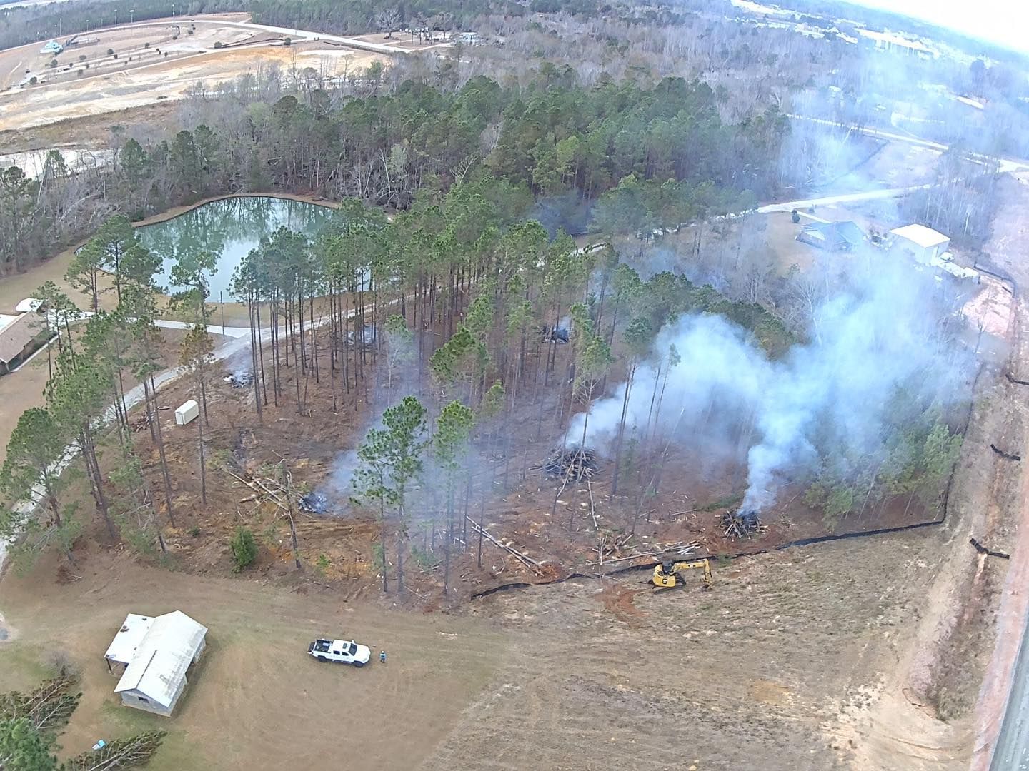 Aerial view of a clearing in a wooded area with burning debris, a pond, and heavy machinery.