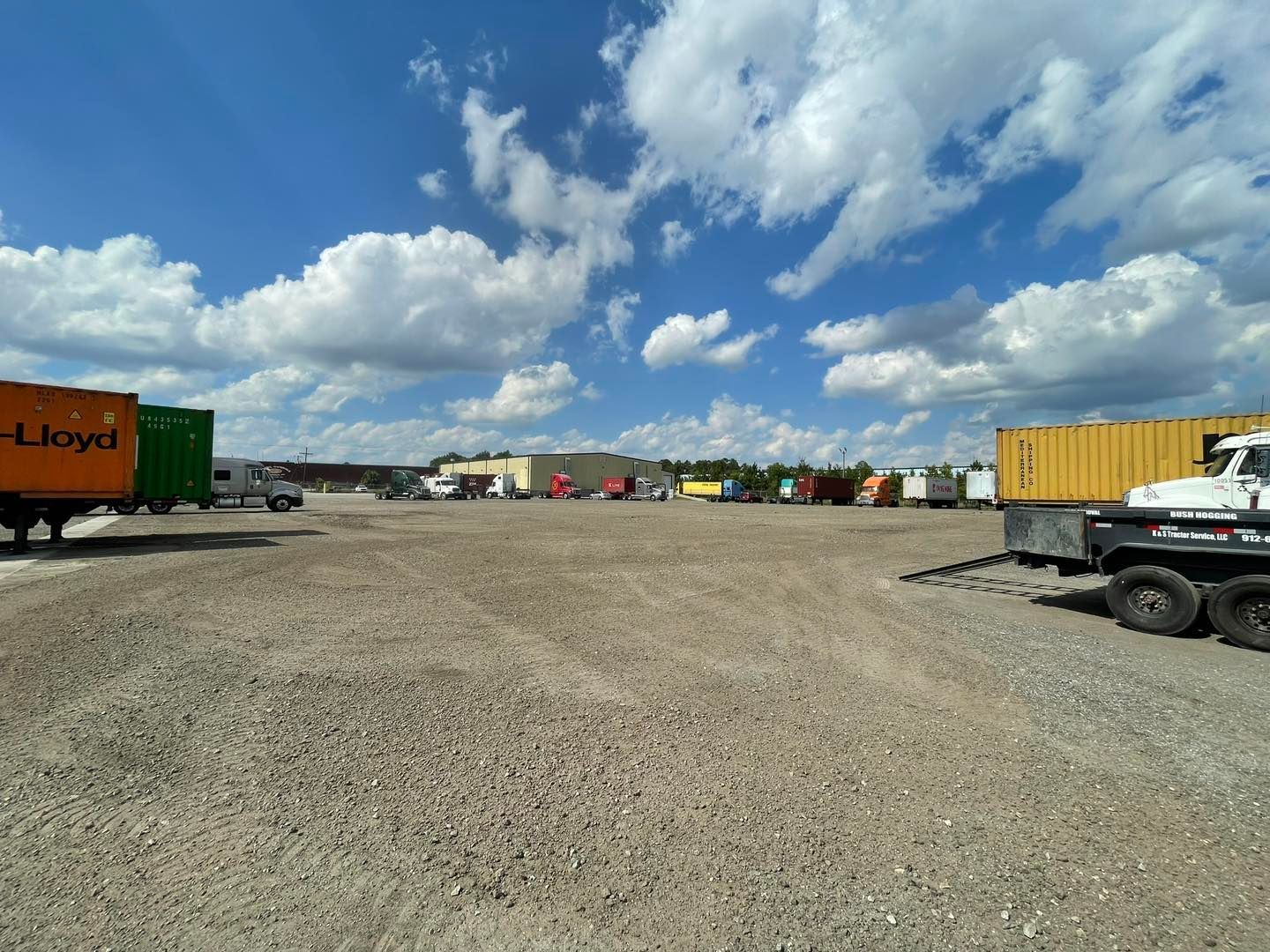 Gravel lot with several semi-trucks and shipping containers under a blue sky with clouds.