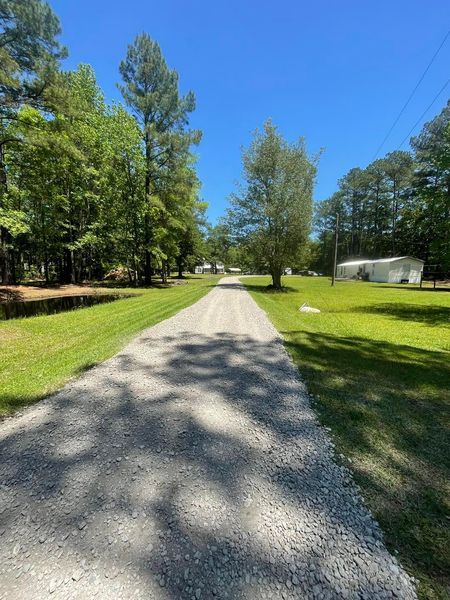 Gravel road leading through grassy area with trees and buildings on a sunny day.