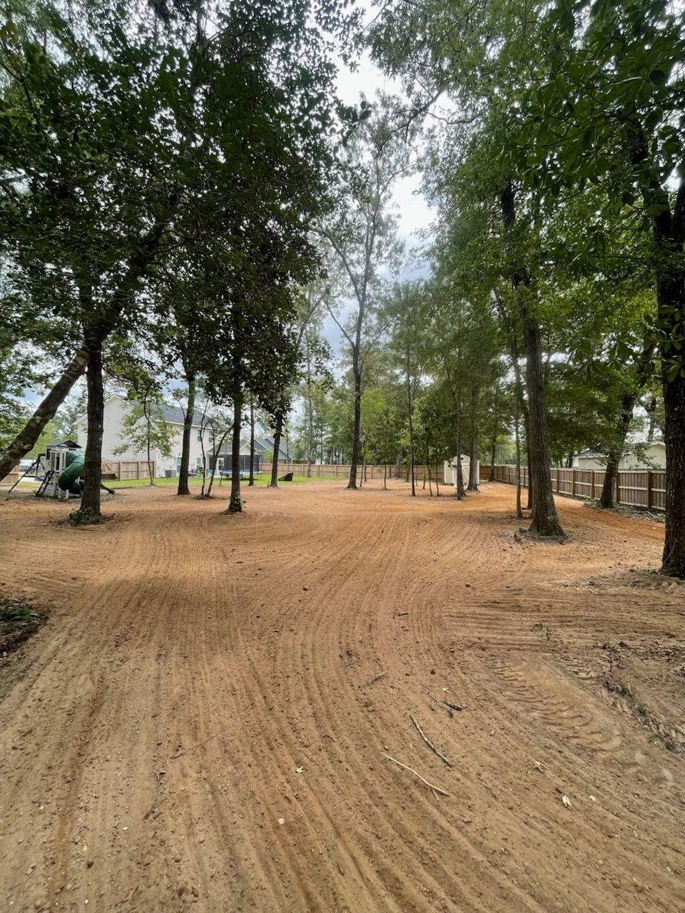 A dirt lot with tire tracks, surrounded by trees. Houses and a cloudy sky are in the background.