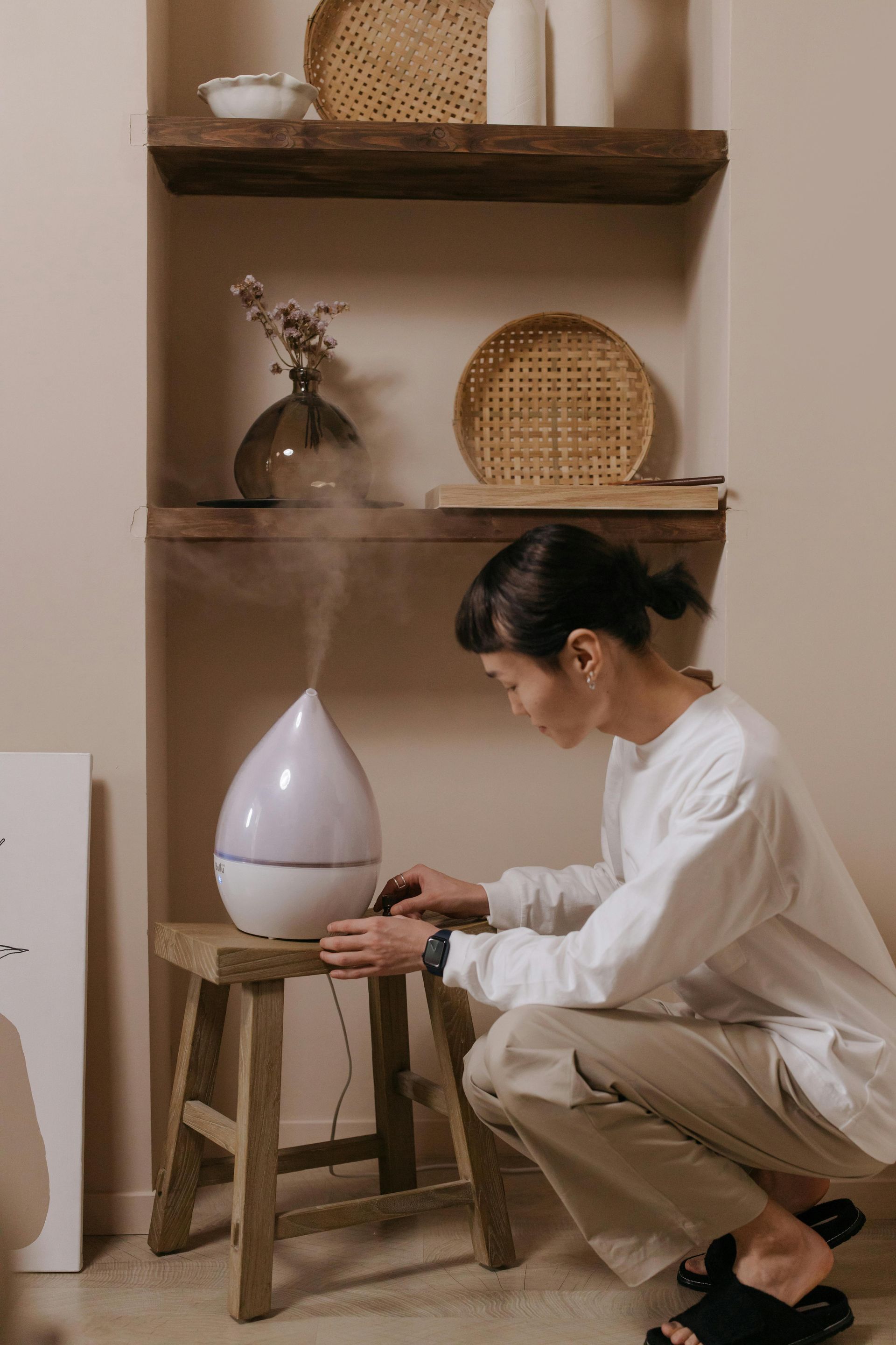 A woman is kneeling down next to a humidifier on a stool.
