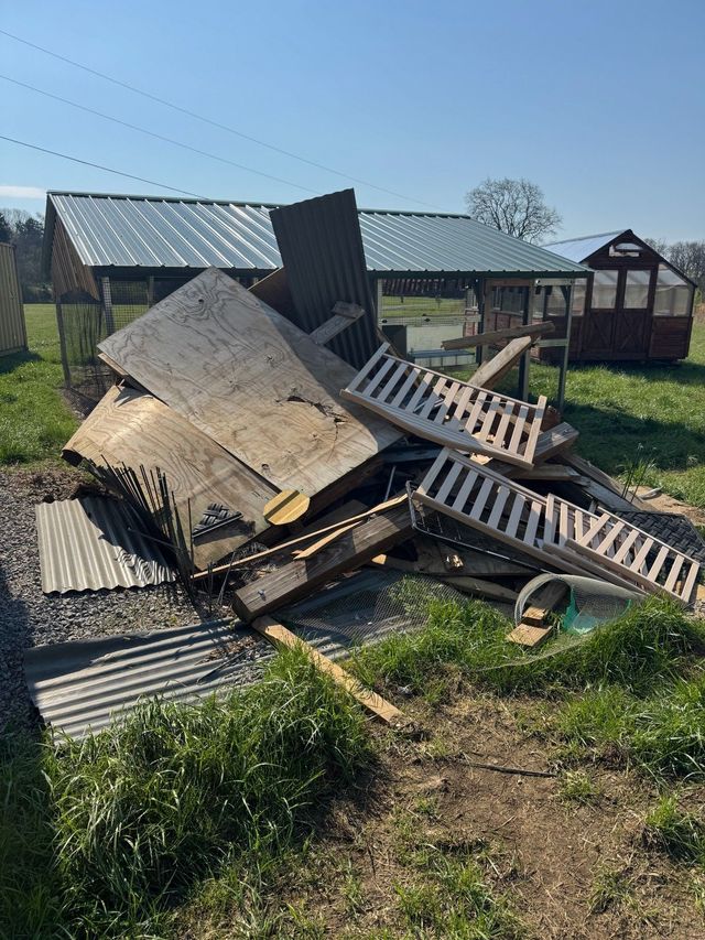 Collapsed wooden structure and debris piled in a grassy yard beside a house on a sunny day
