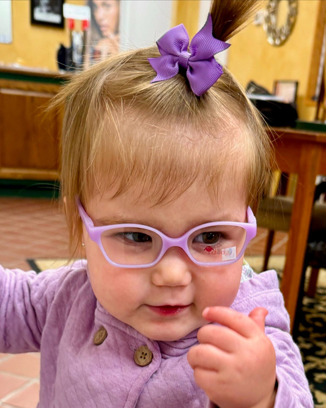 Toddler with light purple glasses and bow, wearing a lavender top, in a room.