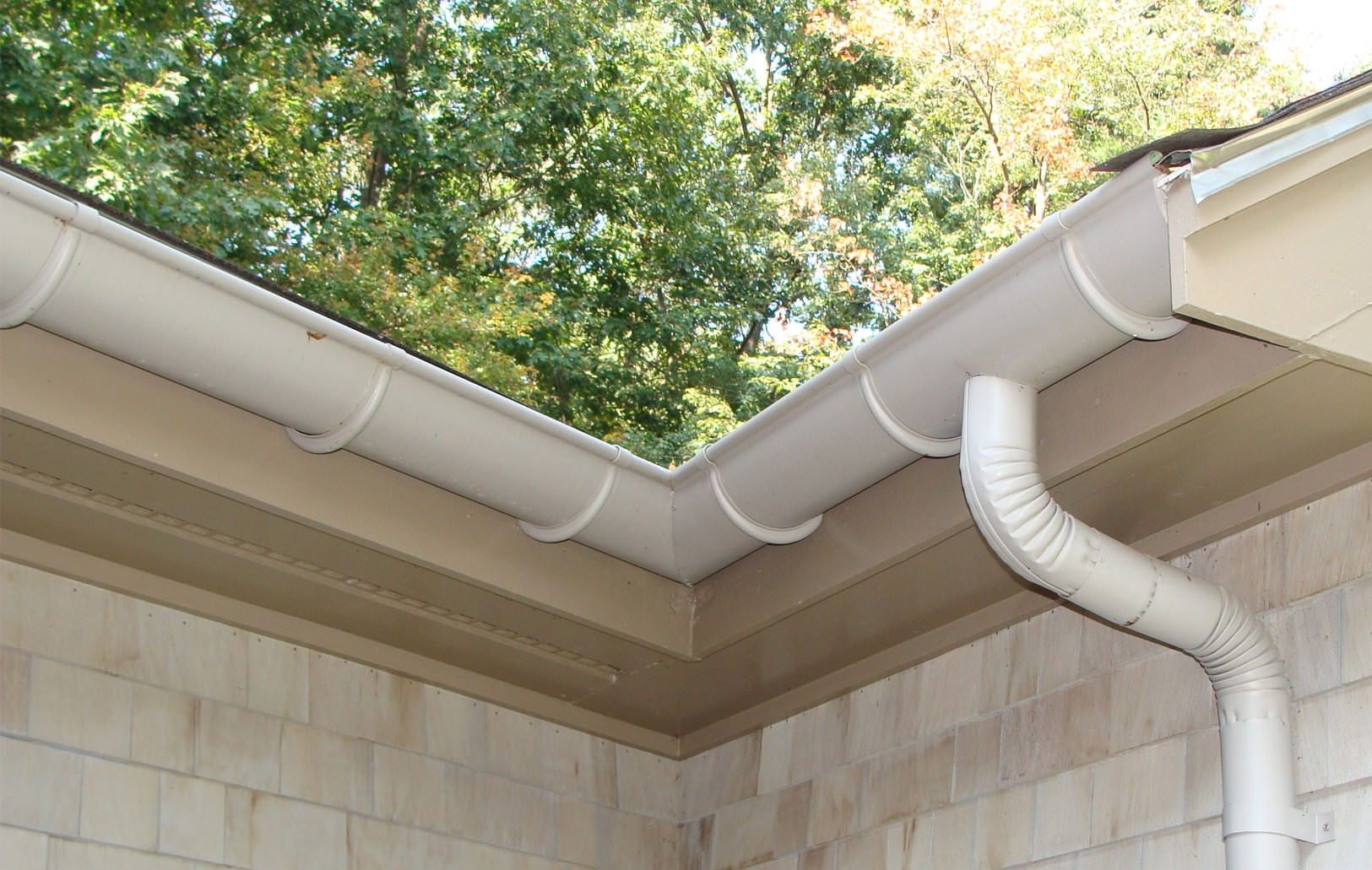 Beige gutter system on a house corner, with a downspout and trees in the background.