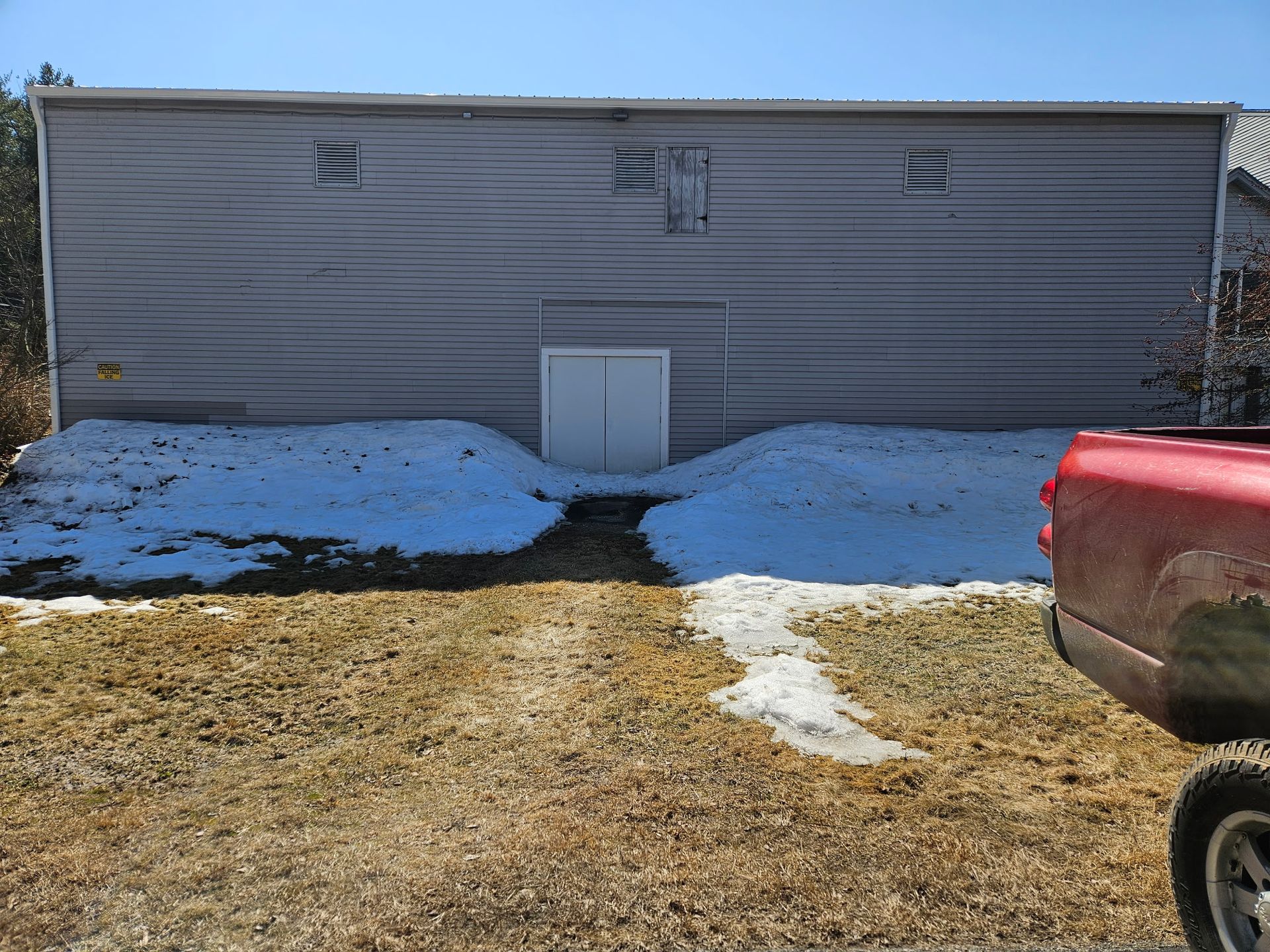 Gray building with snow drifts, doorway, and red pickup truck.