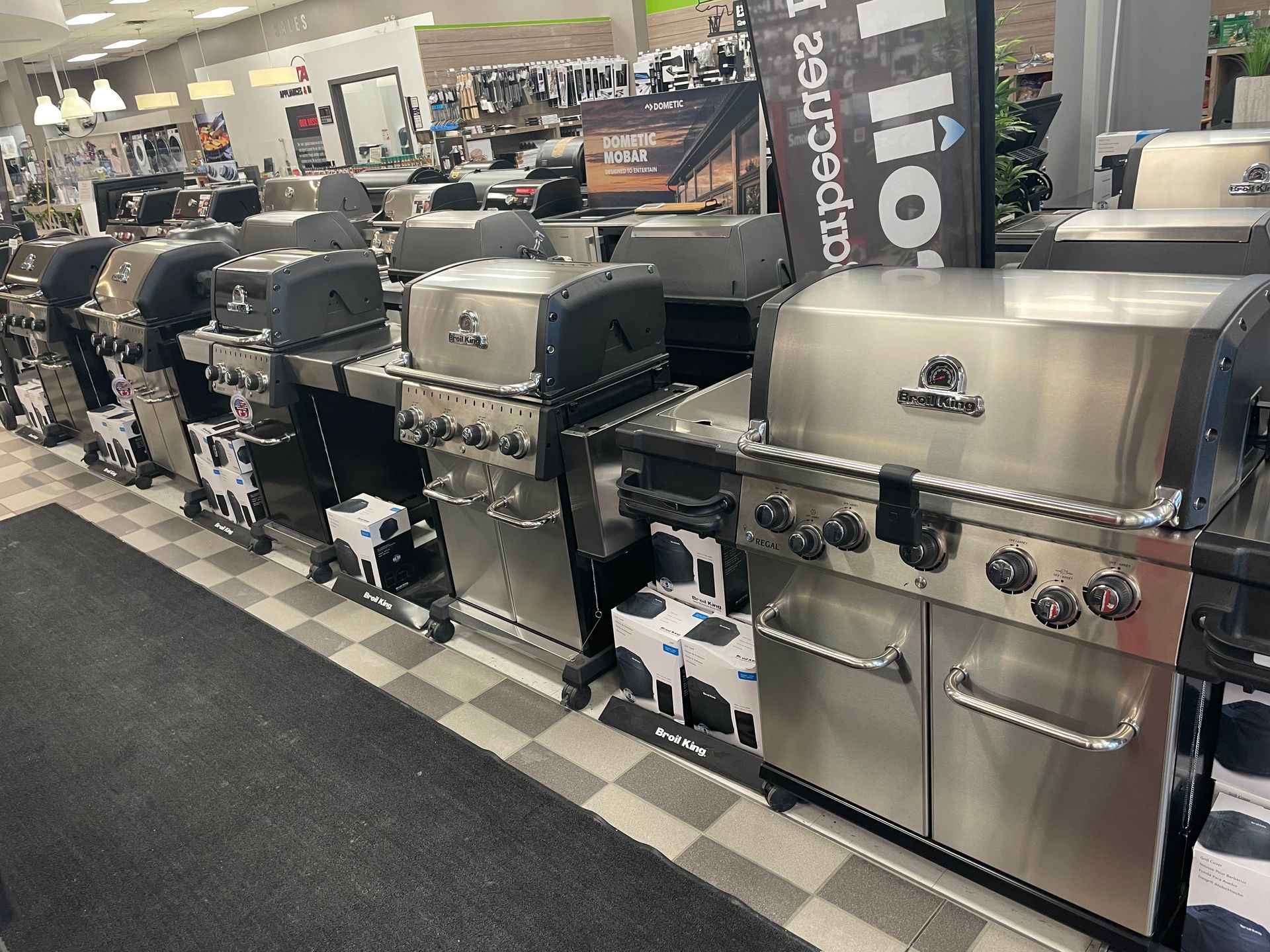Row of outdoor grills on display in a showroom, including stainless steel and black models on a checkered floor.