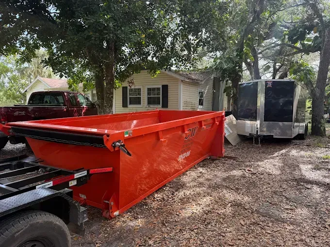 Orange dumpster on trailer parked in front of small house and trailer.
