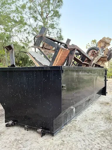 Black dumpster overflowing with metal scrap, sitting outdoors in a yard.