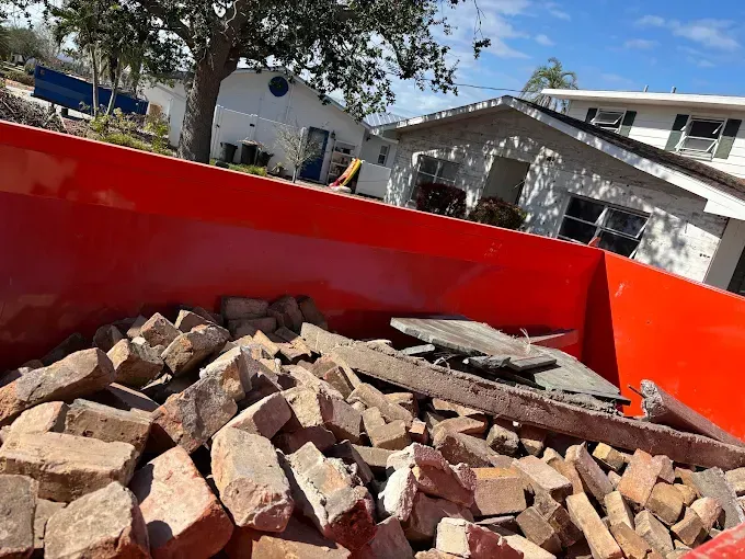 Red dumpster filled with brick debris; houses in the background, likely from demolition or construction.