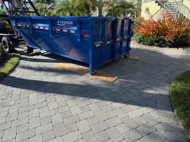 Blue dumpster on brick pavers, being delivered by a truck on a sunny day.