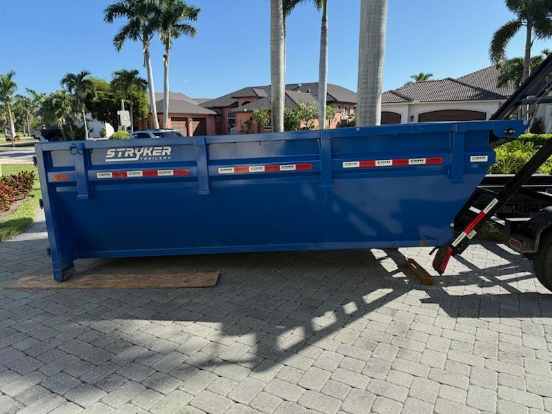 Blue Stryker dumpster on a trailer parked on a paved driveway in front of houses with palm trees.
