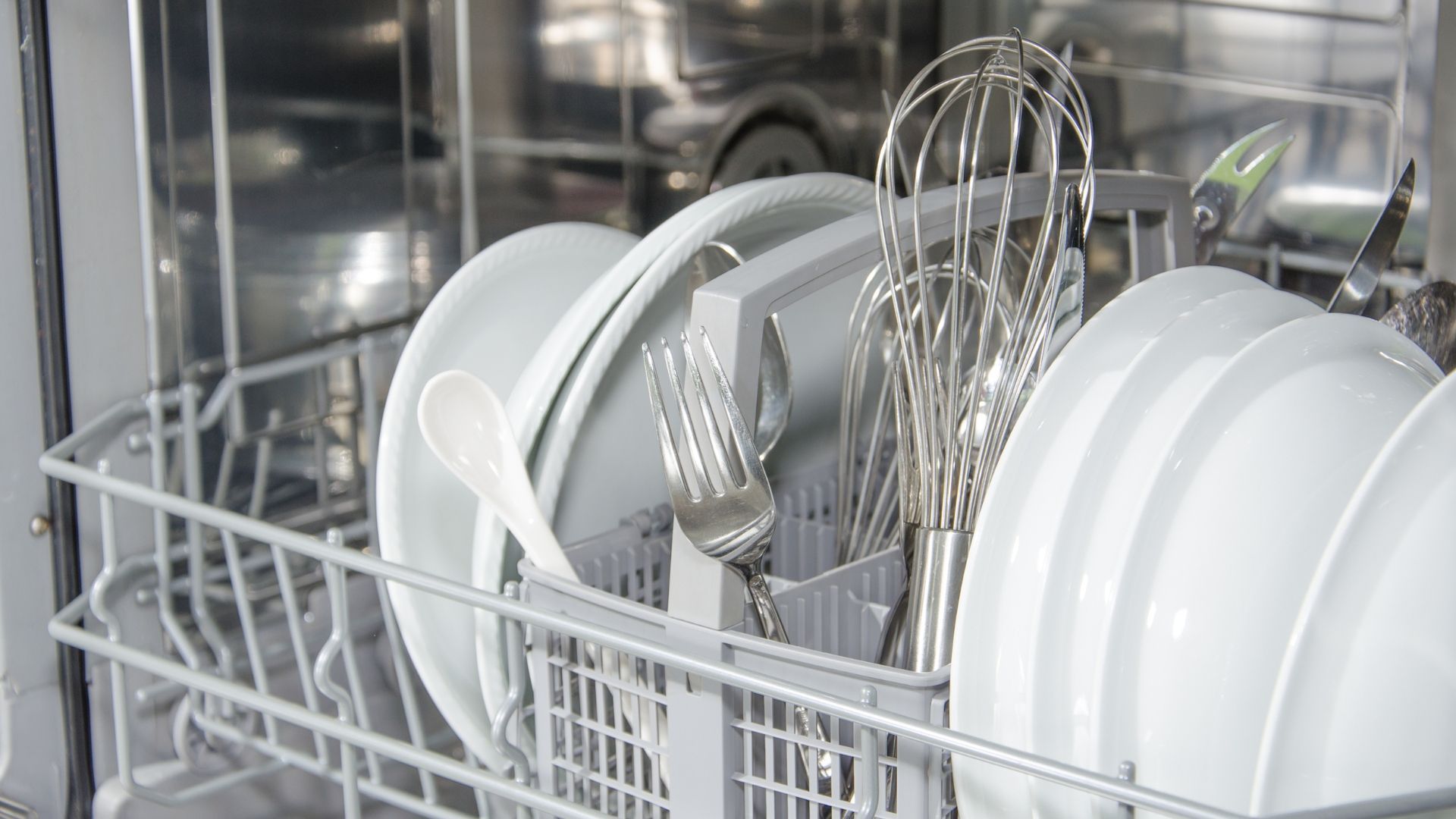 Dishwasher interior with clean white plates, utensils, and whisk in the rack.