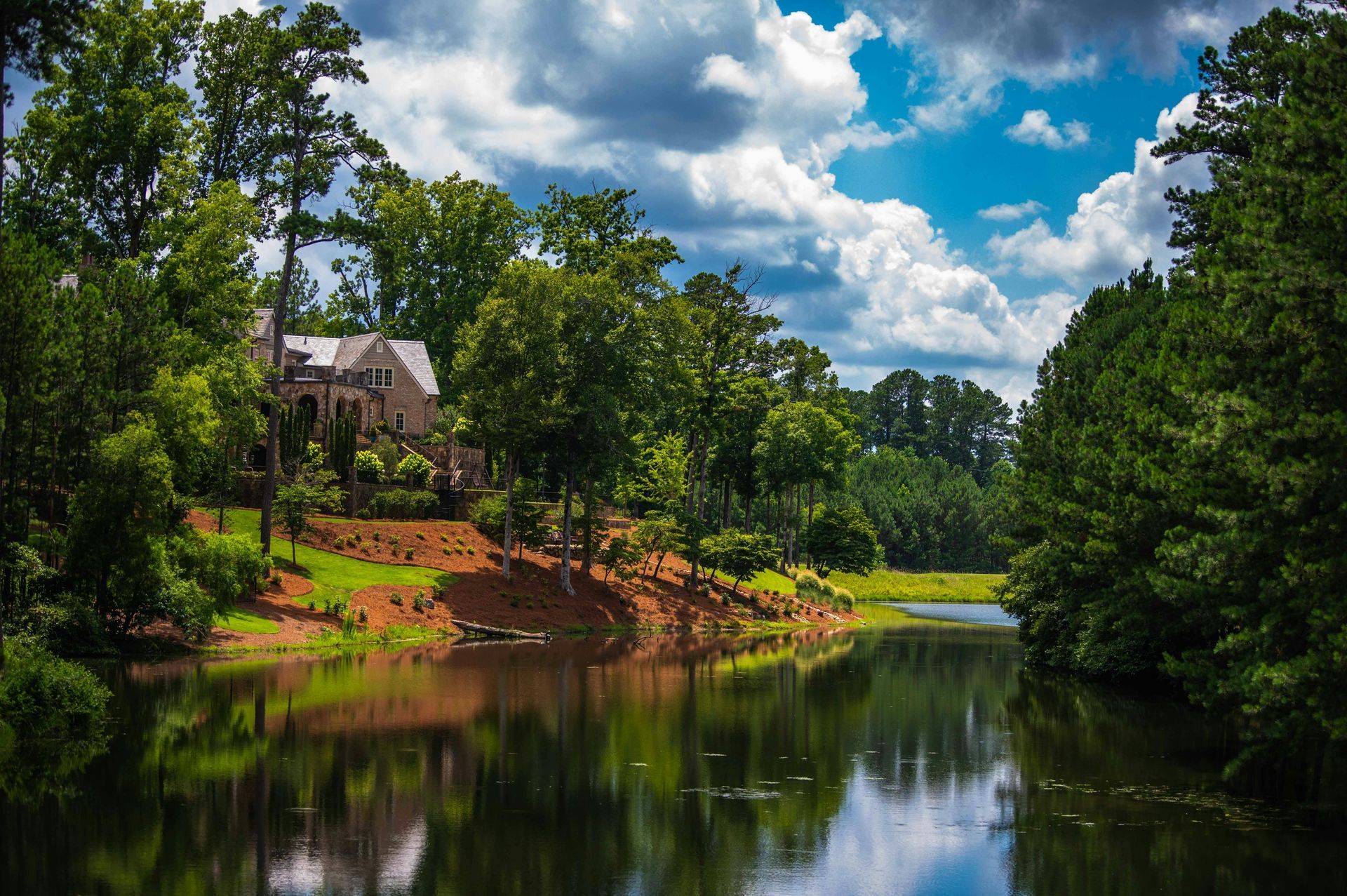 A house nestled on a lake, surrounded by lush trees under a partly cloudy, blue sky.