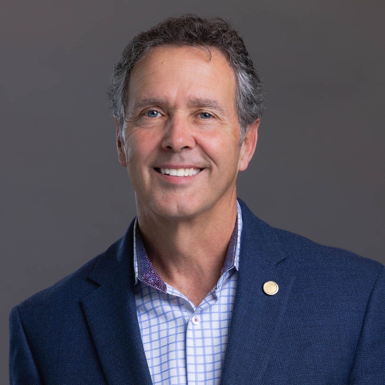 Man in blue blazer, smiling, light blue button-down shirt, gold lapel pin, gray background.