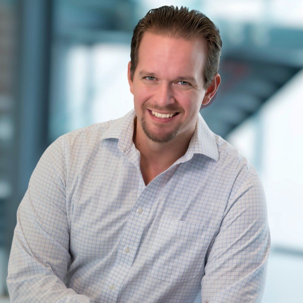 Man smiling, wearing a light blue checked shirt, indoors with a blurred background.