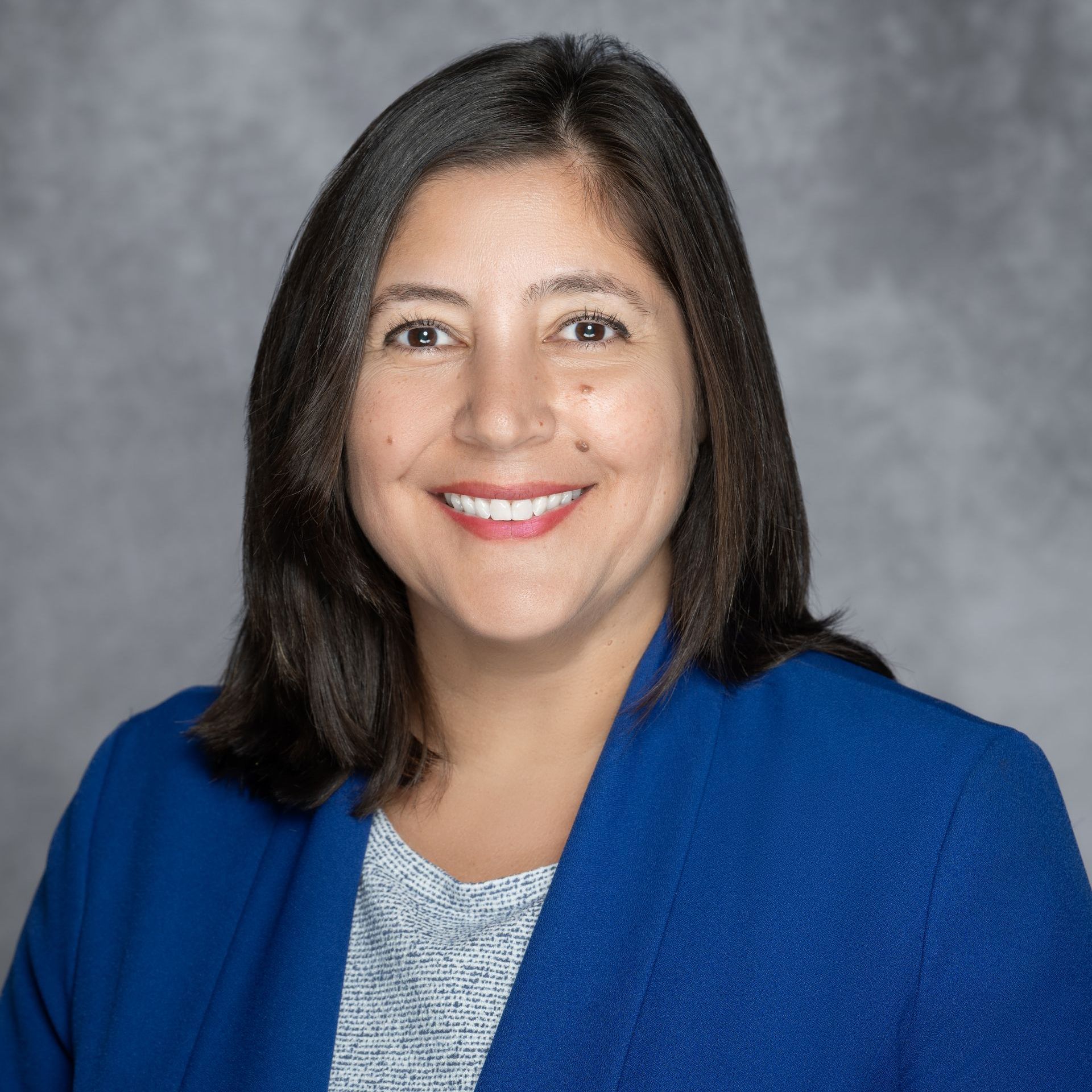 Woman with dark hair smiles, wearing a blue blazer over a gray top, against a gray background.