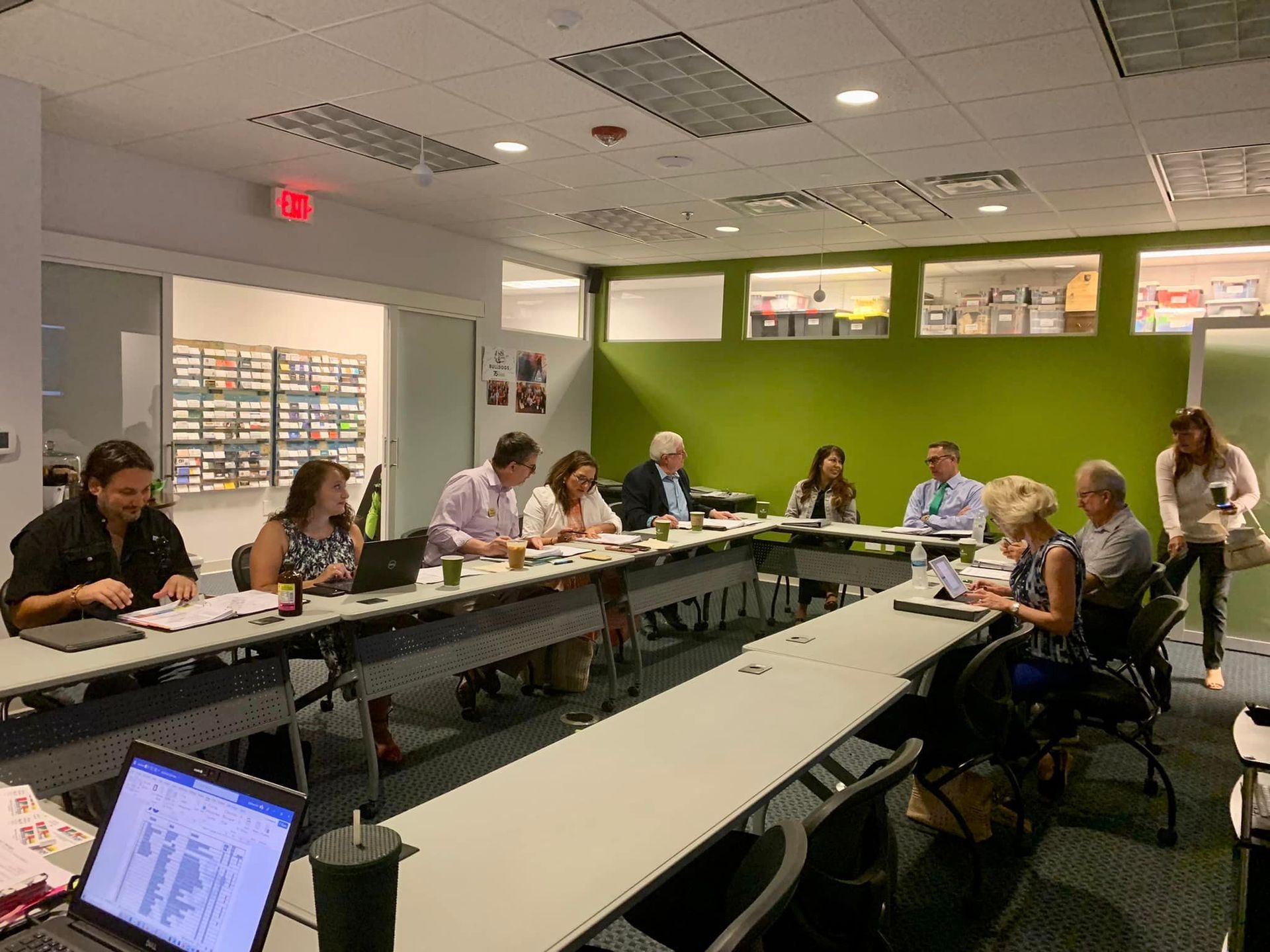 People seated at a table in a meeting room. Some are taking notes, one uses a laptop. Brightly lit room with green and white walls.