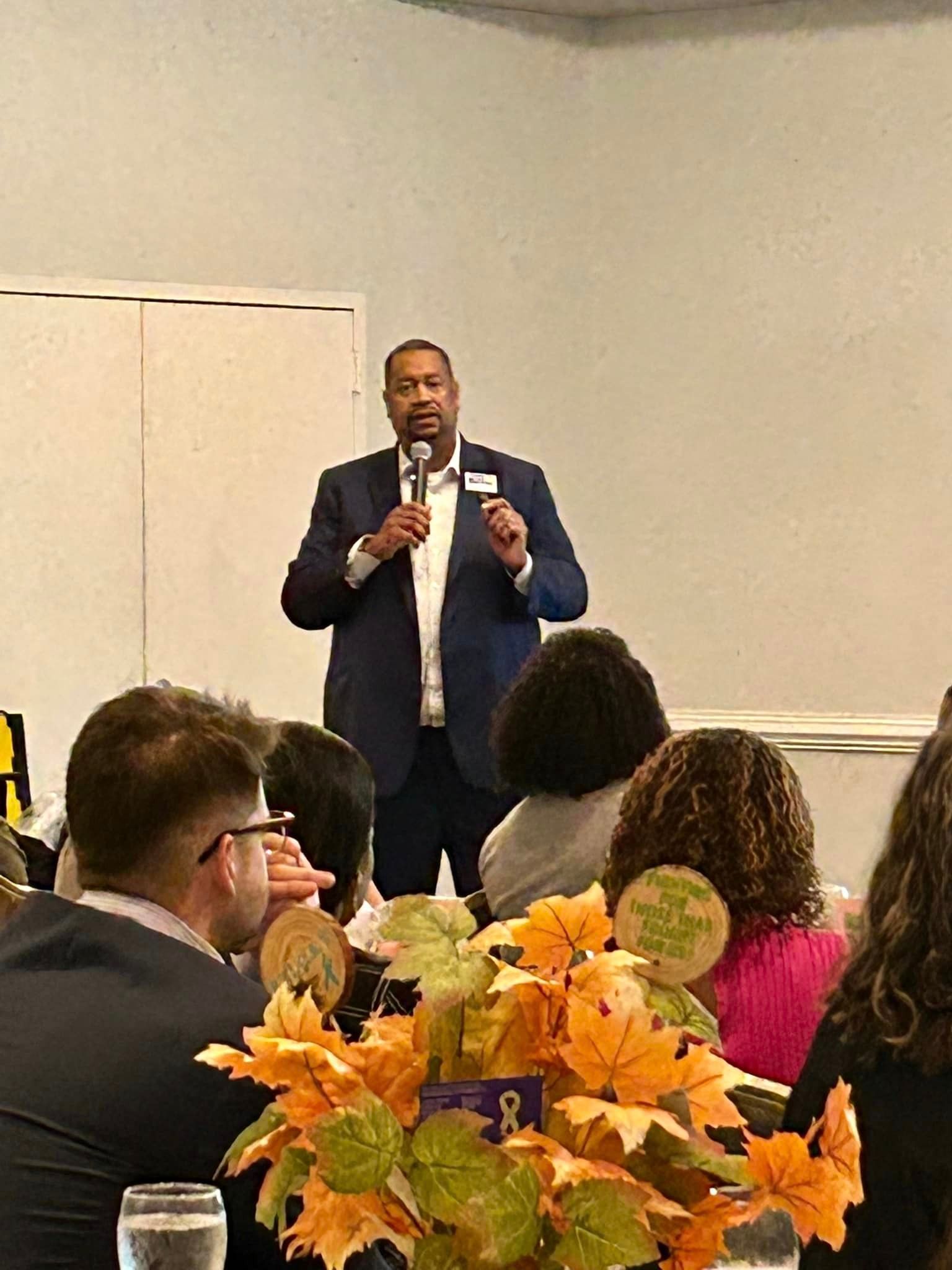 Man in suit speaks into microphone at a gathering. Attendees seated at tables with fall centerpieces.