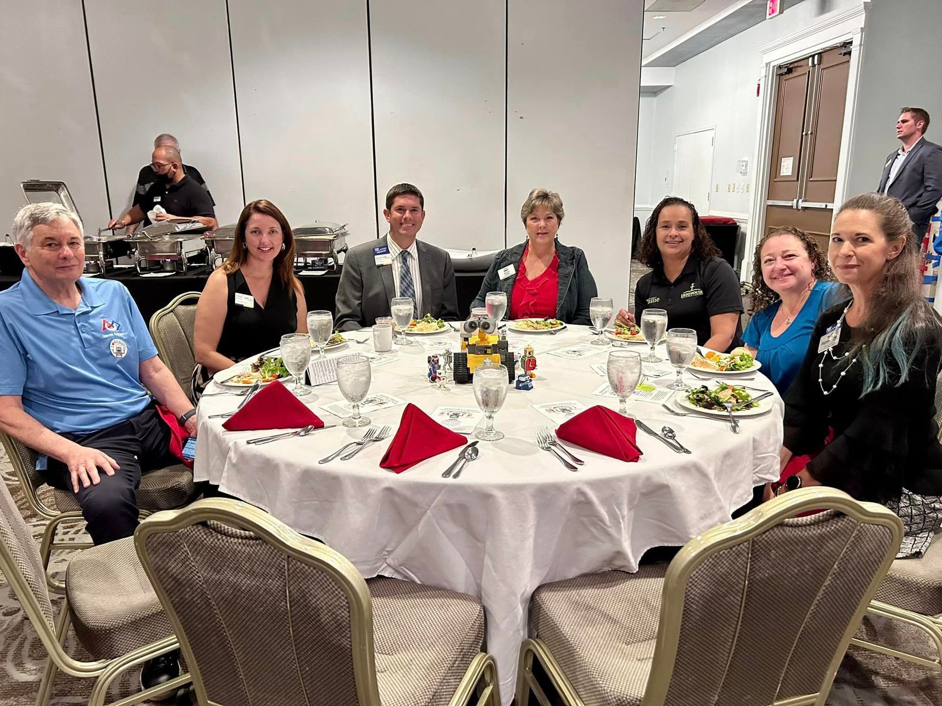 Group of people seated at a round table with red napkins and salad. Buffet in background.