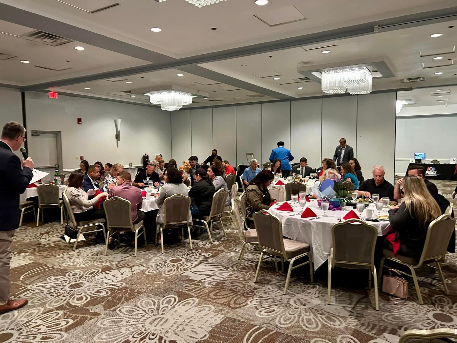 People seated at round tables in a banquet hall, listening to a speaker. White tablecloths, red napkins.
